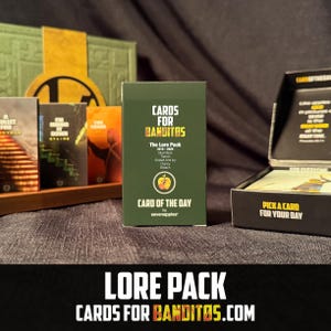 May include: A green card deck box labeled "CARDS FOR BANDITOS" with "The Lore Pack" text. The box features a logo and the text "CARD OF THE DAY." Other card boxes are visible in the background, along with the text "LORE PACK CARDS FOR BANDITOS.COM."