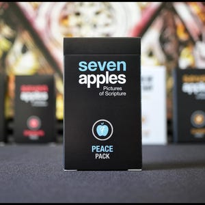 May include: A black box with the words "seven apples" in white and blue lettering. The box also has the words "Pictures of Scripture" in smaller white lettering. The box has a blue apple with a white number 7 inside and the words "PEACE PACK" in white lettering.