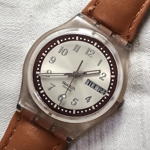 May include: A Swatch Swiss watch with a light-colored face, dark brown ring, and brown leather band. The watch face displays the day and date. The watch has a clear plastic case and a brown leather strap.