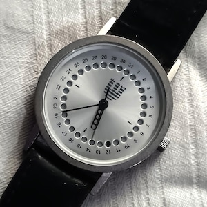 Könnte beinhalten: Silberfarbene Armbanduhr mit schwarzem Lederarmband. Das Zifferblatt hat ein kreisförmiges Design mit Zahlen und Punkten, die das Datum anzeigen. Die Zeiger sind schwarz, und die Uhr hat eine kleine Krone. Zifferblattdurchmesser ca. 4 cm.