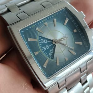 May include: Silver-toned wristwatch with a square face and a matching metal band. The watch face has a blue-green outer ring and a light silver-colored center with the numbers 30, 35, and 40 visible. The watch hands are silver.