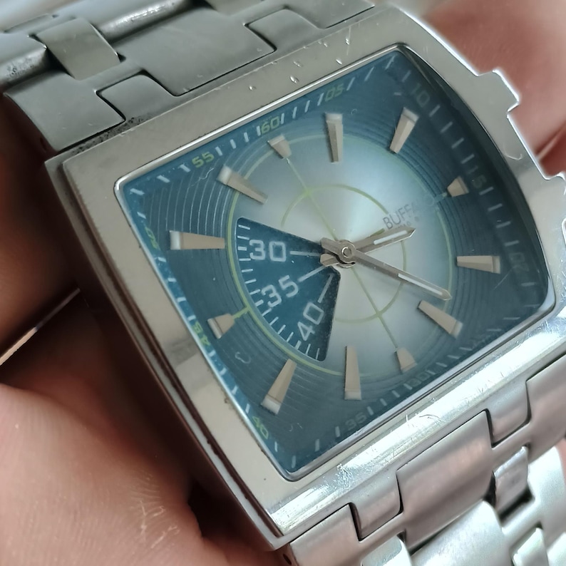 May include: Silver-toned wristwatch with a square face and a matching metal band. The watch face has a blue-green outer ring and a light silver-colored center with the numbers 30, 35, and 40 visible. The watch hands are silver.