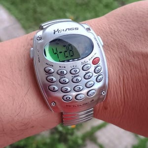 May include: Silver digital calculator watch showing the time 4:28. The watch features a calculator keypad with number and function buttons. The watch strap is silver and the watch is on a wrist.
