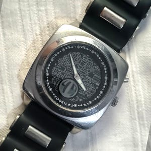 Könnte beinhalten: Eine Fossil-Armbanduhr mit einem quadratischen, silberfarbenen Gehäuse und einem schwarzen Zifferblatt mit detailliertem, weißem Grafikdesign. Die Uhr hat silberne Zeiger und ein schwarzes Armband mit silbernen Akzenten. Das Wort "FOSSIL" ist auf dem Zifferblatt aufgedruckt.