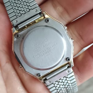 May include: Close-up of a Casio A159W digital watch. The watch has a silver stainless steel band and a gold-toned bezel. The watch back is visible, displaying the Casio logo, model number, and water resistance information.