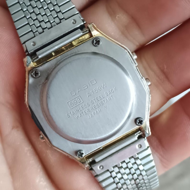 May include: Close-up of a Casio A159W digital watch. The watch has a silver stainless steel band and a gold-toned bezel. The watch back is visible, displaying the Casio logo, model number, and water resistance information.