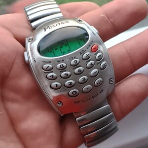 May include: A silver digital calculator watch with a segmented display showing the time. The watch face has a keypad with number and function buttons. The watch band is made of linked metal.