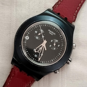 May include: A black Swatch chronograph watch with a black dial, white hands, and a red leather strap. The watch face displays the Swatch Swiss logo and has multiple sub-dials. The watch case is black and has two buttons on the side.