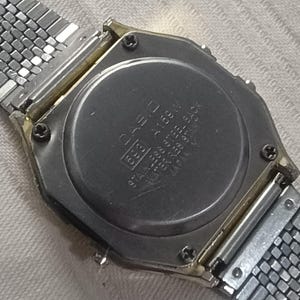 May include: Close-up of the back of a vintage Casio digital watch. The watch has a stainless steel back and a silver metal band. The watch face displays the Casio logo and model information.
