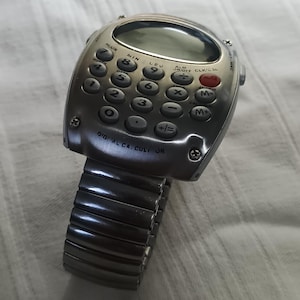 May include: A silver calculator watch with a digital display and a flexible metal strap. The watch face has calculator buttons and the text "DIGITAL CA. CULI OR". This watch combines technology with a retro aesthetic.