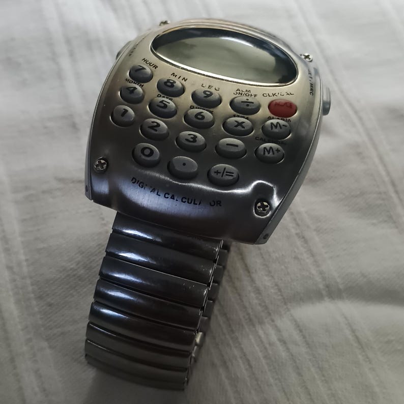 May include: A silver calculator watch with a digital display and a flexible metal strap. The watch face has calculator buttons and the text "DIGITAL CA. CULI OR". This watch combines technology with a retro aesthetic.