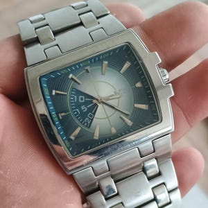May include: Silver-toned BUFFA wristwatch with a rectangular face and a matching metal band. The watch face features a silver and teal dial with gold-colored accents and a date display. The watch is held in a hand.