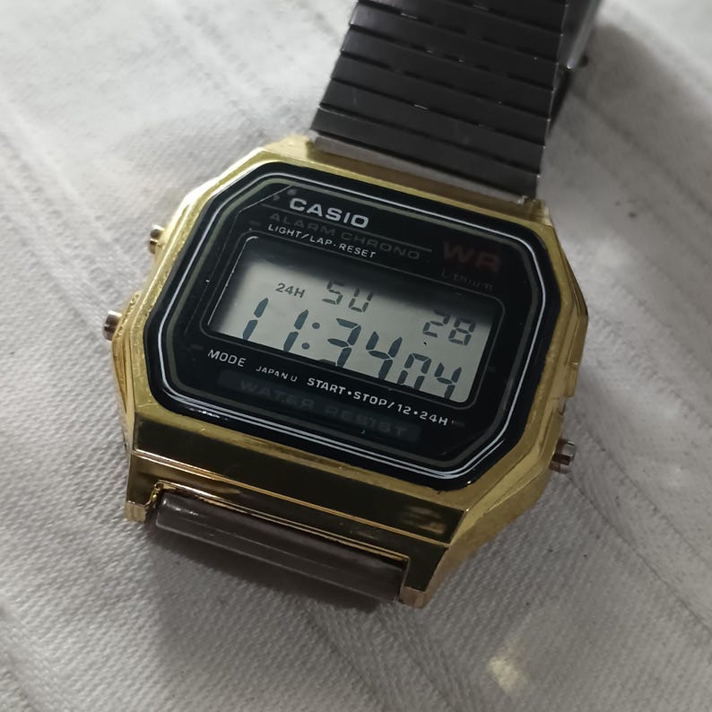 May include: A gold-toned Casio digital watch with a black face displaying the time as 11:34:04. The watch has a black metal band and features the Casio logo and other text on the face. The watch is water resistant.