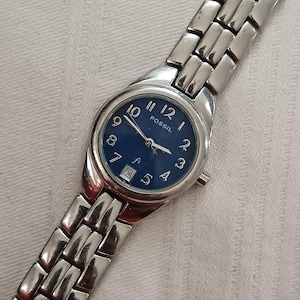 Könnte beinhalten: Eine silberfarbene Fossil-Uhr mit blauem Zifferblatt und Datumsanzeige. Die Uhr hat ein silbernes Armband und ein rundes Zifferblatt mit weißen Ziffern und Zeigern. Die Uhr wird auf einer weißen Oberfläche präsentiert.