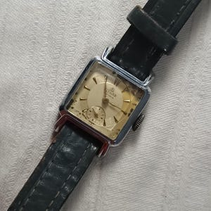 Op de afbeelding: Vintage rechthoekig polshorloge met een zilverkleurige kast, crèmekleurige wijzerplaat en zwarte leren band. De wijzerplaat heeft goudkleurige wijzers en markeringen, met een kleine secondenwijzer. De band is donkergroen.