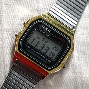 May include: A gold-toned Casio digital watch with a black display showing the time. The watch has a red accent and a silver metal band. The display reads 11:33:19. The watch face includes the text "CASIO", "WR", and "WATER RESIST".