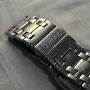 May include: Close-up of a silver-toned watch band clasp. The clasp is engraved with the word "BUFFALO" and the words "DAVID BITTON". The watch band is made of interlocking rectangular links.