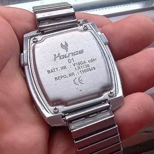 May include: A silver digital watch with a rectangular face and a metal strap. The watch back shows the brand name "Youngs", model number "01", and battery details. The strap is a flexible, expandable design.