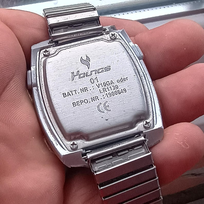 May include: A silver digital watch with a rectangular face and a metal strap. The watch back shows the brand name "Youngs", model number "01", and battery details. The strap is a flexible, expandable design.