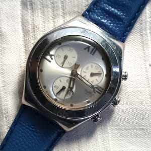 May include: Silver-toned Swatch chronograph watch with a light gray face and blue leather strap. The watch features three sub-dials and Roman numeral hour markers. The blue strap is textured.