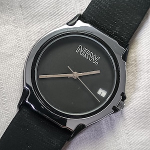 May include: A black wristwatch with a silver-colored bezel and a black leather band. The watch face is dark with the letters "NRW." and a small date window. The hands are silver-colored.