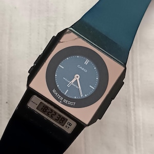 May include: A black Casio digital watch with a square face and a blue dial. The watch features a digital display showing the time, and the words "WATER RESIST" and "ILLUMINATOR" are visible. The watch band is black and blue.