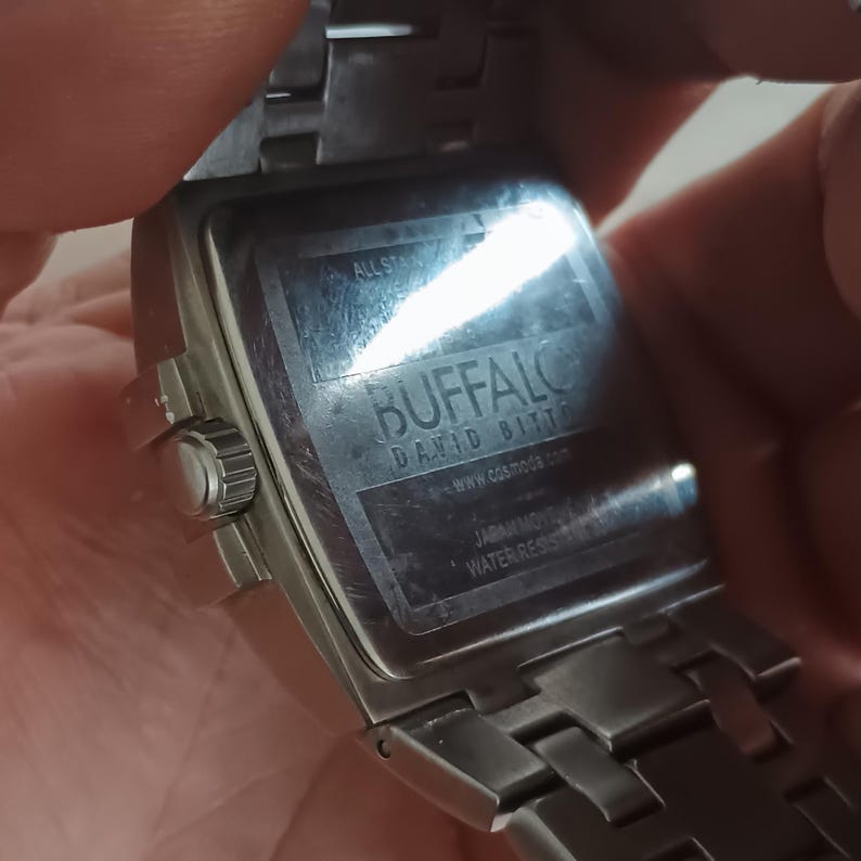 May include: A stainless steel watch with a rectangular face. The watch has a silver band and a silver face. The back of the watch has the brand name "BUFFALO DAVID BITTON" and the text "JAPAN MOVT WATER RESISTANT".