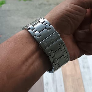 May include: A silver-toned Buffalo David Bitton watch band is clasped on a wrist. The band is made of rectangular metal links. The brand name is printed in white on the clasp. The watch band is a stylish accessory.