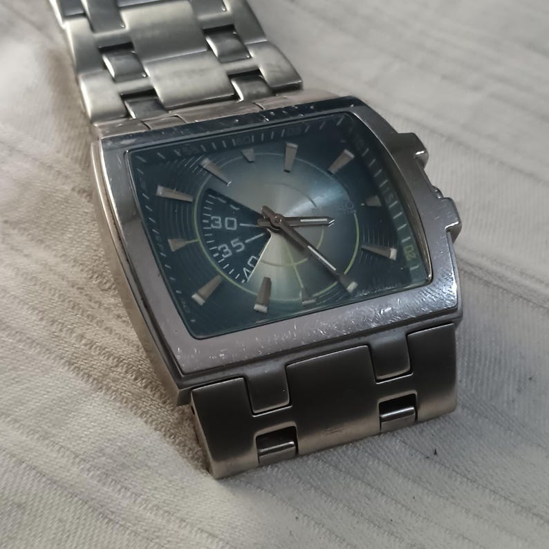 May include: A silver-toned wristwatch with a square face and a matching metal band. The watch face is a gradient of blue and silver, with silver hands and markers. The watch displays the time.