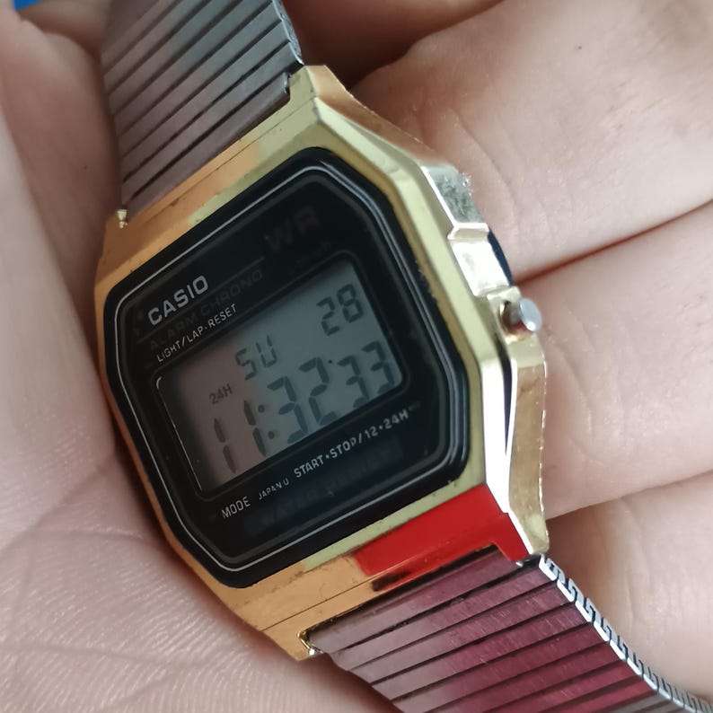May include: A gold-toned Casio digital watch with a black face displaying the time, date, and day. The watch has a silver mesh band and red accents. The watch face reads "CASIO" and "WR". The time displayed is 11:32:33.
