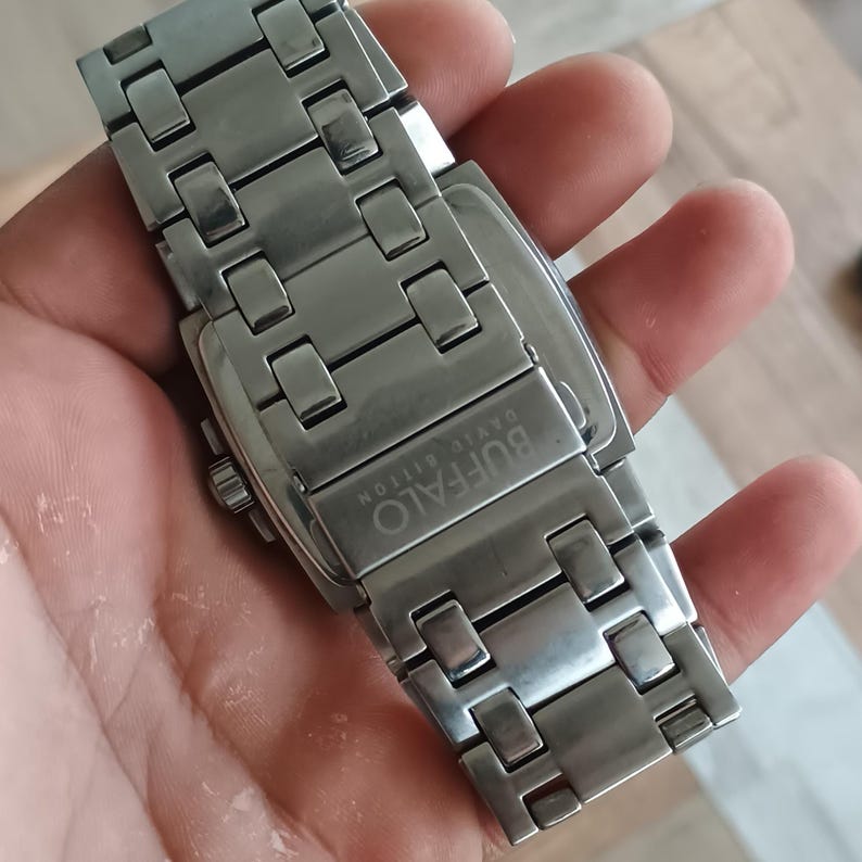 May include: A silver-toned BUFFALO DAVID BITTON wristwatch with a rectangular face and a stainless steel band. The watch band is composed of rectangular links. The watch is held in a hand.