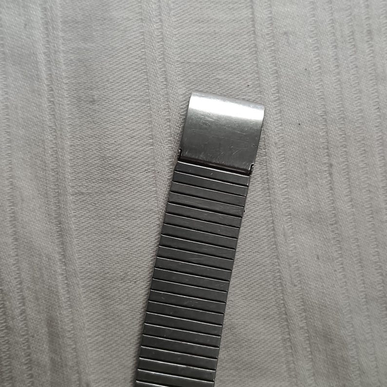 May include: A silver-toned watch band with a rectangular clasp and a flexible, expandable design. The band is composed of numerous small, rectangular metal links, creating a textured appearance. The watch band is shown against a light gray background.