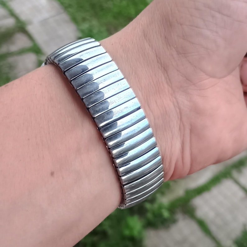 May include: A silver-coloured, expandable watch strap is shown on a wrist. The strap is made of multiple, curved, rectangular links that are connected. The links are shiny and reflect light. The strap is flexible and stretches to fit the wrist.