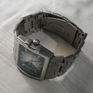 May include: A silver-toned wristwatch with a rectangular face and a matching metal band. The watch face is a dark teal color with a lighter design in the center. The band is made of rectangular links.