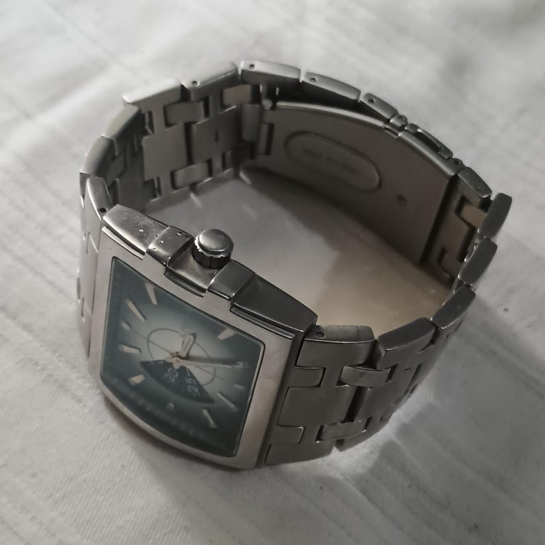May include: A silver-toned wristwatch with a rectangular face and a matching metal band. The watch face is a dark teal color with a lighter design in the center. The band is made of rectangular links.