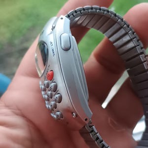 May include: A silver digital wristwatch with a calculator keypad and a flexible metal band. The watch face has a small screen and several buttons, including "JET, MRC", "ALARM", and "CALENDAR".