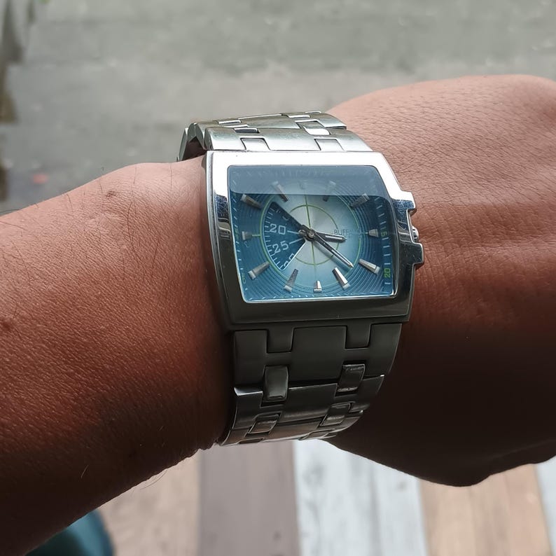 May include: A silver-toned wristwatch with a square face and a blue dial. The watch has a stainless steel band and a unique design. The watch face displays numbers and hands for timekeeping. The watch is worn on a wrist.