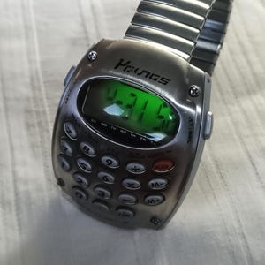 May include: A silver calculator watch with a digital display showing the time. The watch has a full keypad and a metal band. The brand name "Prunas" is visible on the watch face. The time displayed is 4:31.