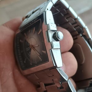 May include: A stainless steel wristwatch with a rectangular face and a silver band. The watch face is dark with silver hands and markers. The watch is held in a hand, showing the side with the crown and band clasp.