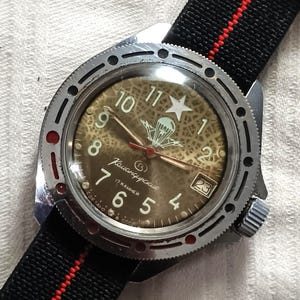 May include: A vintage wristwatch with a brown face, white numerals, and a star at the 12 o'clock position. The watch has a silver bezel and a black and red striped nylon strap. The watch face includes the text "Командирские".