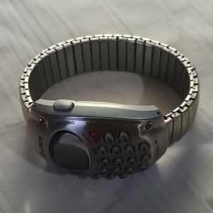 May include: A silver wristwatch featuring a keypad and a small screen. The watch band is made of linked metal segments. The watch face has buttons and a small display screen. The watch has the word "SOURCE" printed on the side.