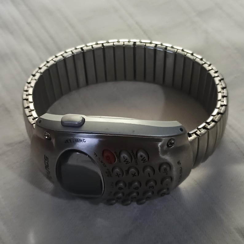 May include: A silver wristwatch featuring a keypad and a small screen. The watch band is made of linked metal segments. The watch face has buttons and a small display screen. The watch has the word "SOURCE" printed on the side.
