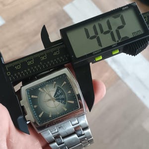 May include: A silver wristwatch with a rectangular face is measured with a digital caliper. The watch face is dark green with silver accents and a circular design. The caliper displays a measurement of 44.2 mm, which is approximately 1.74 inches.