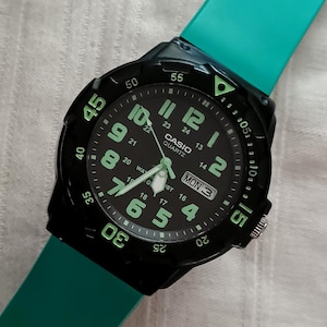May include: Black Casio Quartz watch with a teal band. The watch face is black with large, luminous green numerals and hands. The day and date are displayed. The watch is set to Monday, the 3rd.
