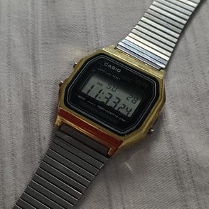 May include: A gold-toned Casio digital watch with a silver metal band. The watch face displays the time as 11:33:24. The watch has a black digital display with the Casio logo and other text.