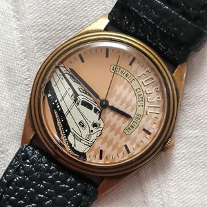 May include: A Fossil watch with a black leather strap. The watch face is a light peach colour with a black and white train graphic. The watch has gold-coloured trim and the words "AUTHENTIC CLASSIC ORIGINAL FOSSIL" around the face.