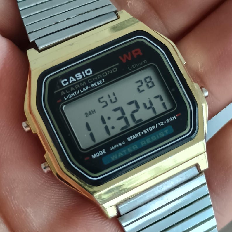 May include: A gold-toned Casio digital watch with a silver band. The watch face displays the time, date, and various functions. The watch has a black and gold bezel with the Casio logo and other text.