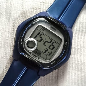 May include: A blue digital wristwatch with a rectangular display showing the time 15:26 and the date. The watch has a blue strap and several buttons. The watch face includes the text "CASIO" and "ILLUMINATOR".