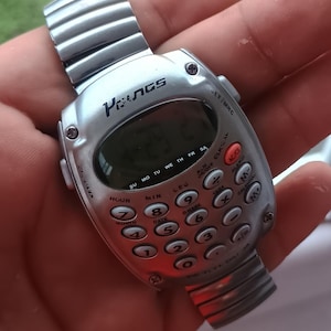 May include: Silver calculator watch with a digital display showing the time. The watch has a metal strap and a keypad with numbers and function buttons. The brand name "Penas" is visible on the watch face.