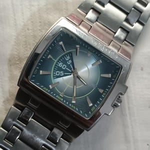May include: A silver-toned wristwatch with a rectangular face and a matching metal band. The watch face is a gradient of blue and silver, with white numerals and hands. The watch has a sporty, modern design.